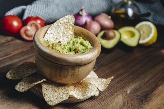 delicious guacamole in a wood surrounded with nachos next to an avocado and other ingredients, typical mexican healthy vegan cuisine with rustic dark food photo style
