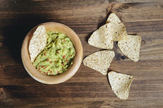 top view of traditional guacamole with nachos in a wood bowl on wooden table, typical mexican healthy vegan cuisine with rustic dark food photo style