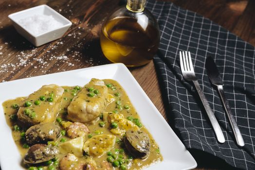 healthy traditional spanish hake dish in green seafood sauce with clams, prawns, green peas and egg, typical mediterranean cuisine with rustic dark food photo style