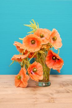 Vase of casually arranged pale pink poppies on a wooden table against a turquoise background