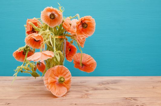 Numerous beautiful cut pink poppies with long winding stems, overflow from a glass vase on a wooden table, against a painted turquoise background
