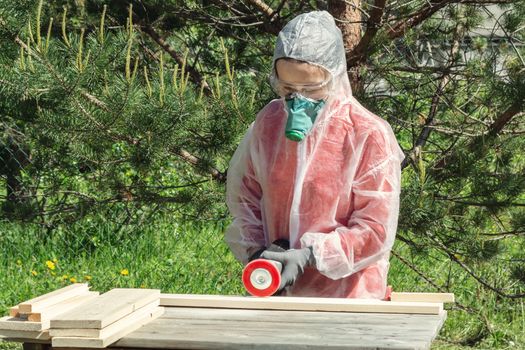 Woman carpenter in respirator, goggles and overalls handles a wooden board with a Angle grinder.