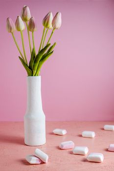 Copyspace with colorful mini marshmallows on a table next to pink tulips in a white vase on a pink background