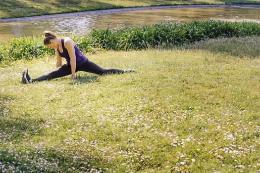 young sports woman doing flexibility exercises and stretching legs on the grass outdoors in the park, modern healthy lifestyle and sport concept, copy space for text
