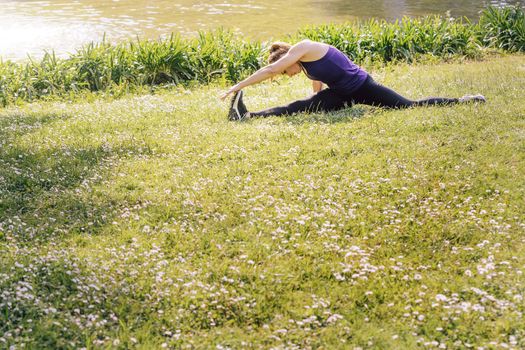 young sporty woman doing flexibility exercises and stretching legs on the grass outdoors in the park, modern healthy lifestyle and sport concept, copy space for text