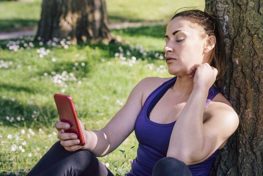 sports woman looking at her phone while resting after exercise sitting next to a tree, healthy modern lifestyle and sport concept