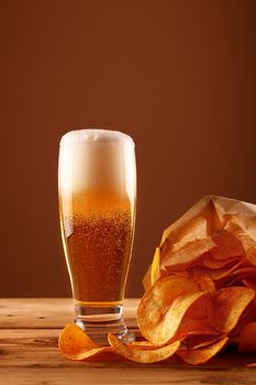 Close up one glass of lager beer with white froth and bubbles and paper bag of potato chips on wooden table over dark brown background with copy space, low angle side view