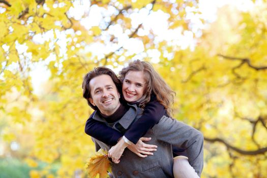 Smiling mid adult couple hug in autumn forest of yellow maple trees giving piggyback ride