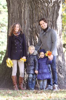 Happy smiling family of parents and children in autumn park standing near big tree trunk