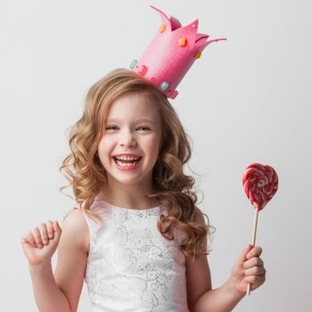 Beautiful little candy princess girl in crown holding big pink heart shaped lollipop and smiling