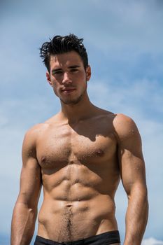 Young fit athletic man at beach in summer day showing muscular torso, looking at camera