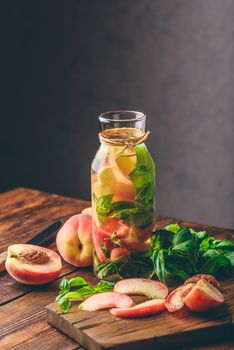 Bottle of Detox Water with Sliced Peach and Basil Leaves. Knife and Ingredients on Cutting Board. Vertical Orientation.