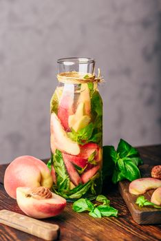 Bottle of Infused Water with Sliced Peach and Basil Leaves. Knife and Ingredients on Cutting Board. Vertical Orientation.