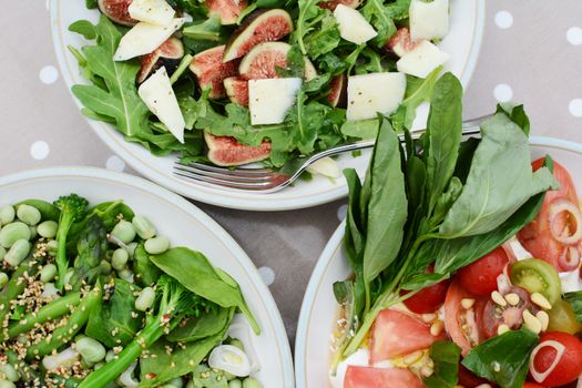 Delicious fresh salads with figs, tomatoes, spinach, broad beans and broccolini seen on a table from above