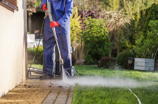 Caucasian Men and the Spring Maintenance and Cleaning. Washing House Backyard Brick Paths Using Pressure Washer. House Surroundings Maintenance. Housing Theme.

