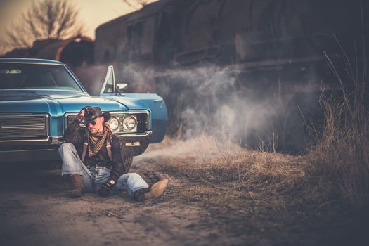 The American Cowboy Story. Caucasian Men Wearing Western Style Clothes and Cowboy Hat, Taking a Moment in Front of His American Classic Muscle Car. Aged and Rusty Steam Locomotive in the Background.