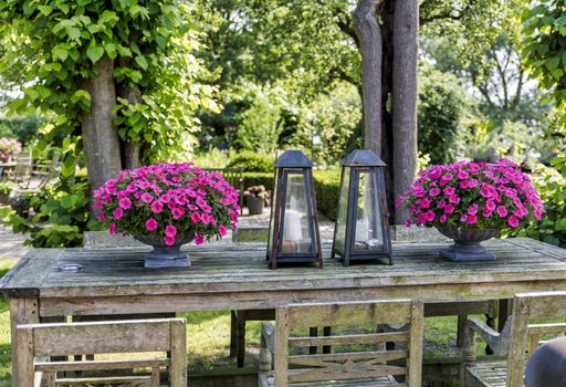 wooden table with chairs with vases with petunias and beautiful old metal lamps in an English garden with greenery and trees