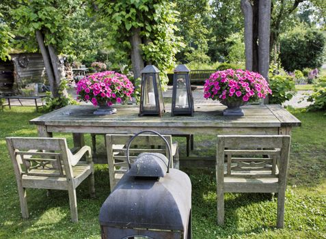 wooden table with chairs with vases with petunias and beautiful old metal lamps in an English garden with greenery and trees