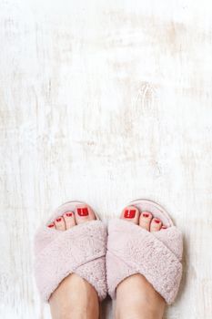 female legs with red nails in home fur fluffy pink slippers on a light wooden background. flat lay. Top view. The concept of a cozy bright girl house.