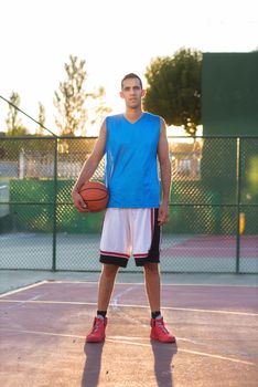 Athlete man holding basketball ball standing on playground at sunset