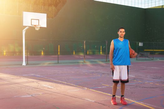 Athlete man holding basketball ball standing on playground at sunset