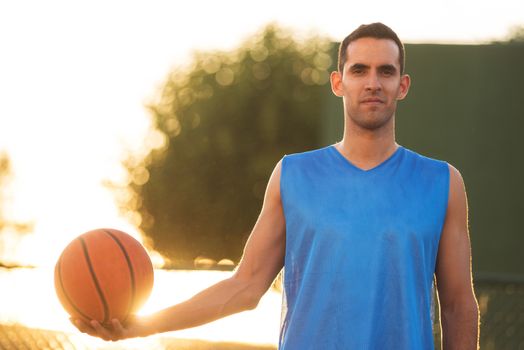 Athlete man holding basketball ball standing on playground at sunset