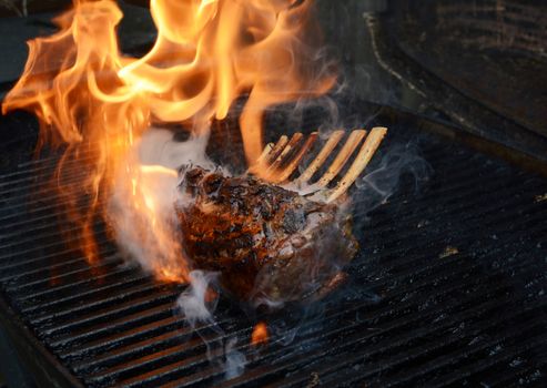 Tender rack of lamb being grilled, with large flames and smoke rising from the barbecue