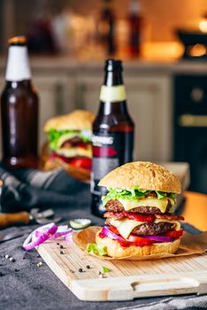 Cheeseburger with Two Beef Patties, Cheddar Cheese, Bacon, Iceberg Lettuce, Sliced Tomatoes and Red Onion. Bottle of Beer and Some Ingredients on Table.