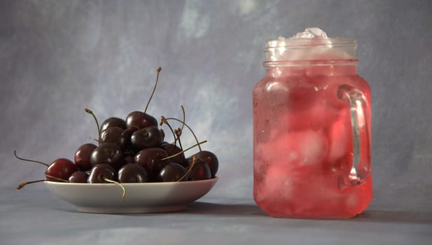 A glass mug with a refreshing juice with ice and a full plate with ripe sweet cherry.
