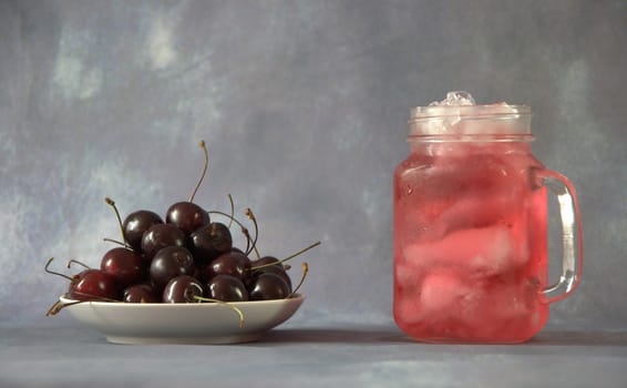 A glass mug with a refreshing juice with ice and a full plate with ripe sweet cherry.