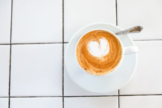 Cappuccino cup on tiled white table background. Foam is decorated with cinnamon heart. Copy space. Top view, located at side of frame. Horizontal. Mock up for social media, food blog, lifestyle.