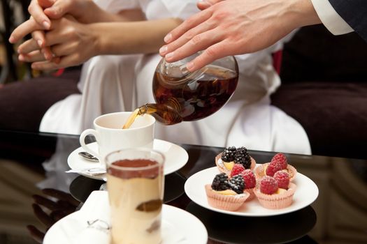 A man cares for a woman: pours her green tea. On the table are desserts: tiramisu and pastries with fresh berries. Without faces, in the frame of the hand. Closeup.