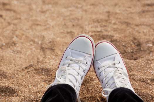 View of the legs in white sneakers on background of shell beach. Atmospheric, melancholic mood, blurred background. For design mockup, content for social media. Horizontal. Close-up