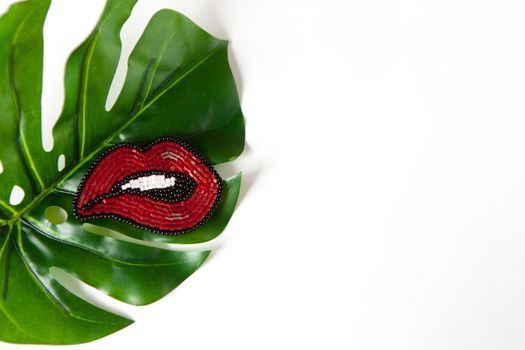 Fashionable brooch in the shape of lips from Japanese beads on green leaf of Monstera on white background. Close-up, flat lay, copy space. Concept trends, fashion, style