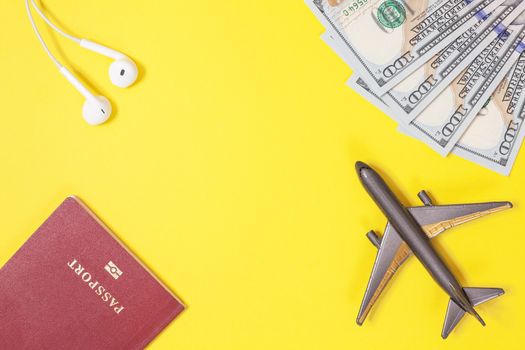 One hundred dollar bills, airplane, headphones, foreign passport on bright yellow paper background. Copy space. Travel and budget trip concept, flat lay. Hand luggage, minimalism, frame of objects