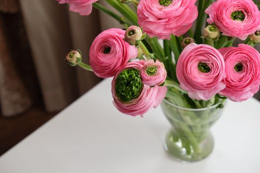 Pink Ranunculus bouquet in transparent glass vase on white table. Close-up. For flower delivery, social media. Soft selective focus. Copy space. Horizontal.