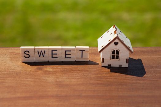 Word sweet of wooden letters and small toy wooden house on brown surface, green grass in background. Concept sweet home, mortgage, dream house, real estate acquisition. Soft selective focus.