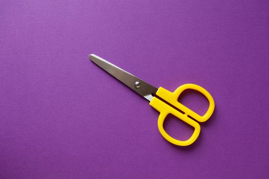 Yellow kid's stationery right-handed scissors on purple papper background. Back to school, DIY concept. Top view, copy space