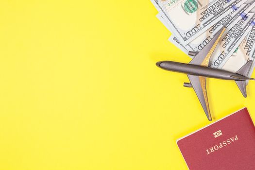 One hundred dollar bills, airplane, headphones, foreign passport on bright yellow paper background. Copy space. Travel and budget trip concept, flat lay. Hand luggage, minimalism, objects in corner