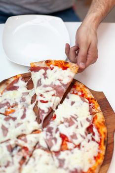 Man took first slice of delicious Italian pizza. In frame hand taking slice of hot pizza with ham on wooden board, white serving plate on white table. Pizza time. Top view