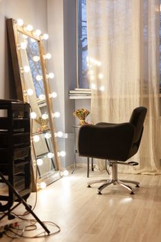 Workplace stylist, hairdresser, makeup artist. Beauty saloon. Black chair for client opposite large floor mirror with bulbs around perimeter, organizer for tools. Evening time, cozy atmosphere