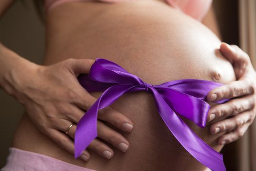 Pregnant woman supports her belly. A purple ribbon is tied to the bow on the belly. Concept of pregnancy as a gift, happiness of parenthood. Lifestyle photo. Horizontal orientation of the frame.
