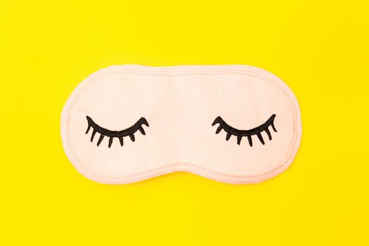 Pastel pink sleep mask with closed eyes embroidered on it with eyelashes on bright yellow neon background. Fashion accessory for sleep. Concept of vivid dreams, accessories for girls and young women