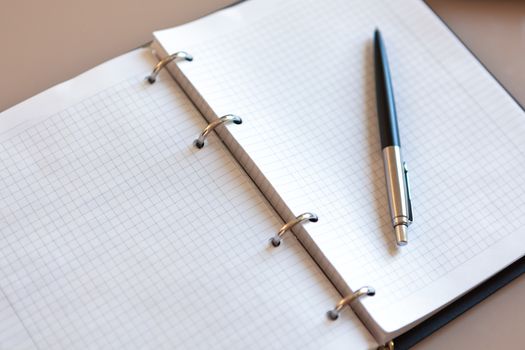 Open notebook with pen lying on it on beige desktop. Notepad sheets on silver brackets, automatic ballpoint pen in silver-black colour. Concept business, work at home. Copy space. Top view, close-up