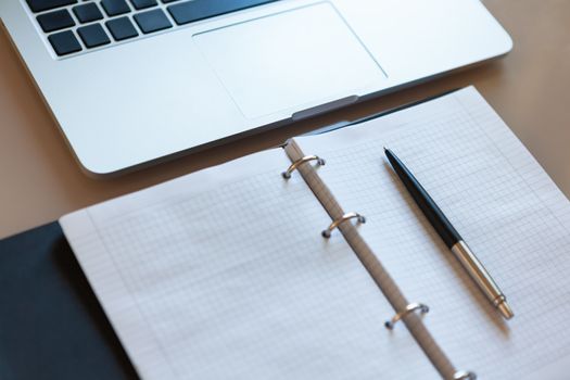 Top view on working space. Laptop and open notebook with pen on beige desktop. Notepad sheets on silver brackets, automatic ballpoint pen in silver-black colour. Business background