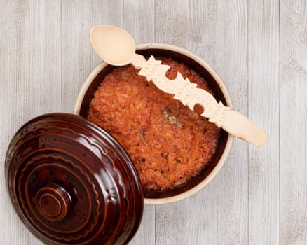 A ceramic pot of traditional Eastern European stewed cabbage and decorated wooden spoon on a white wooden kitchen board.