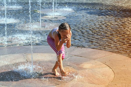 Girl on a sunny summer day are poured water from a fountain.Girl happily in shallow clean water on of city fountain on warm bright summer day.