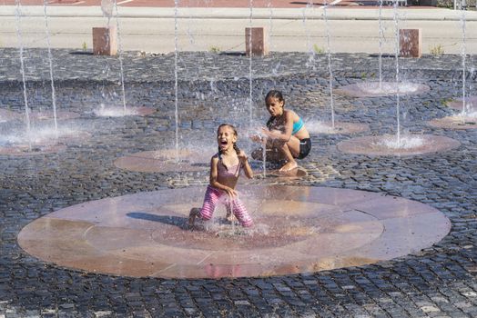 Children on a sunny summer day are poured water from a fountain.Children happily in shallow clean water on of city fountain on warm bright summer day.