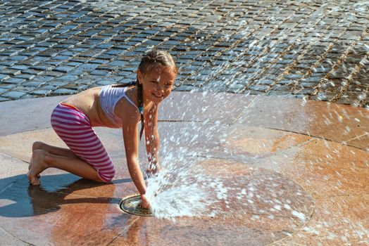 Girl on a sunny summer day are poured water from a fountain.Girl happily in shallow clean water on of city fountain on warm bright summer day.
