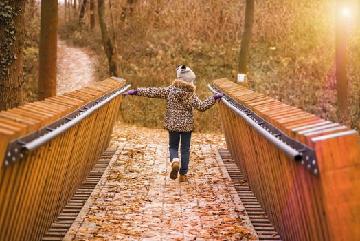 Girl in a coat and hat in an autumn day walks on a wooden bridge. A girl walks across the bridge on an autumn day. An autumn warm sunny day.
